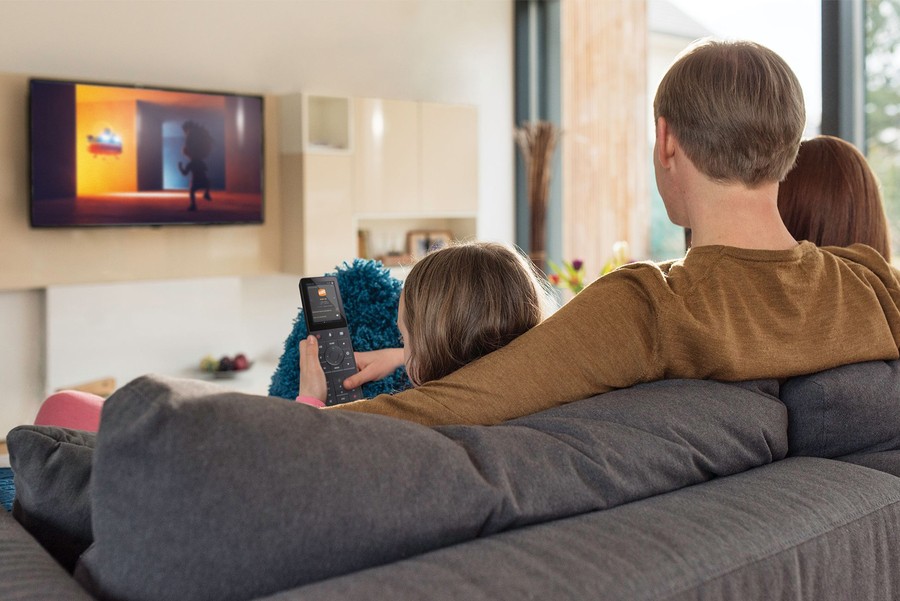 A family sits on a gray sectional in a bright living room watching an animated movie on a wall-mounted TV, with a child holding a smart remote.